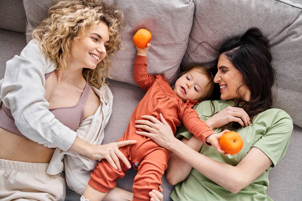 liebevolles lustiges lgbt-Paar auf Sofa liegend mit ihrem kleinen Mädchen, das Mandarinen hält, Familienkonzept - Foto, Bild