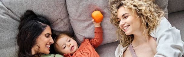 jolly lgbt couple lying on sofa with their cute baby girl holding tangerines, family concept, banner - Photo, Image