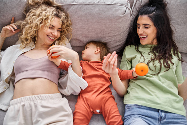 loving happy lgbt couple lying on sofa with their baby girl holding tangerines, family concept - 写真・画像