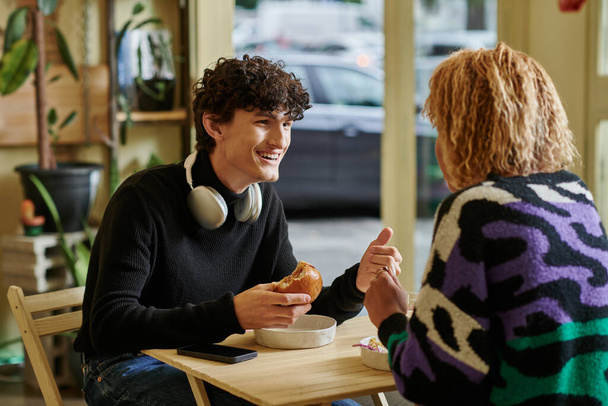 happy curly man with wireless headphones holding tofu burger and chatting with girl in vegan cafe - Photo, Image