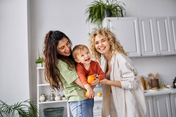 merry lesbian couple having fun together with their cute baby girl holding tangerines, family - Photo, Image