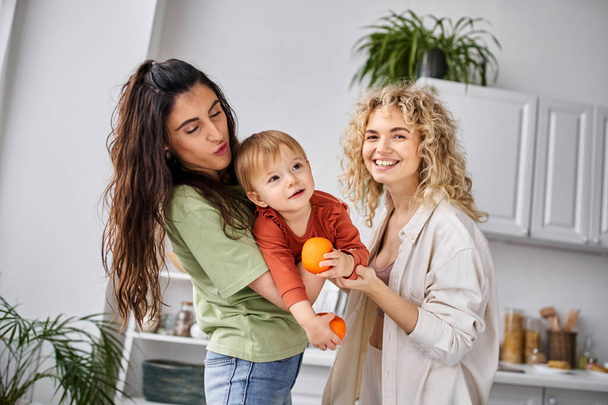 happy pretty lgbt couple having fun together with their cute baby girl holding tangerines, family - Photo, Image