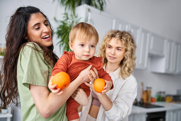 alegre bastante lgbt pareja divertirse juntos con su lindo bebé niña sosteniendo mandarinas, familia - Foto, Imagen