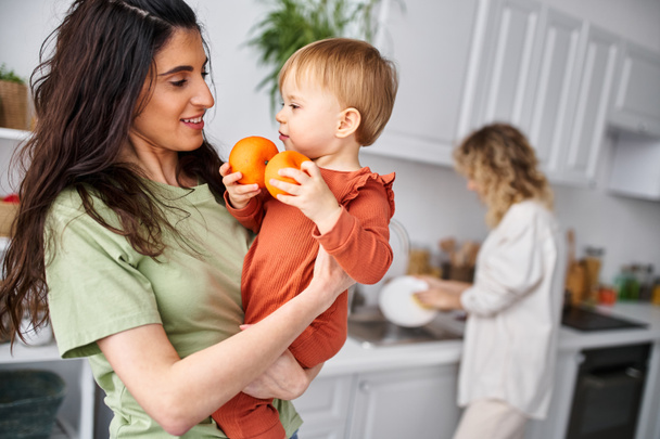 focus op mooie vrouw met dochter houden mandarijnen met haar wazig blond partner op de achtergrond - Foto, afbeelding