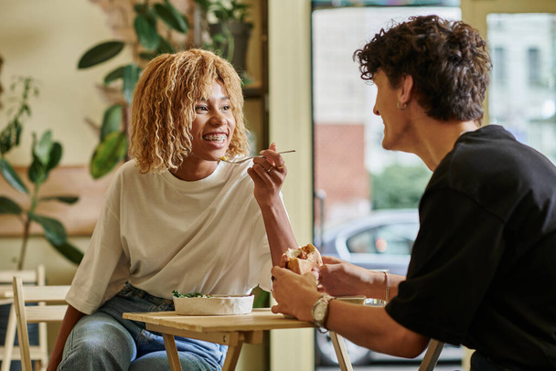 happy african american woman with braces eating salad bowl near blurred boyfriend in vegan cafe - Photo, Image