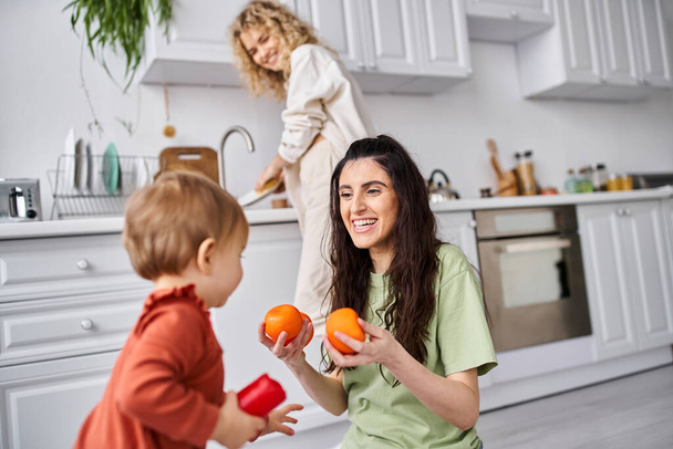 focus on merry woman with daughter holding tangerines with her blurred blonde partner on backdrop - Photo, Image