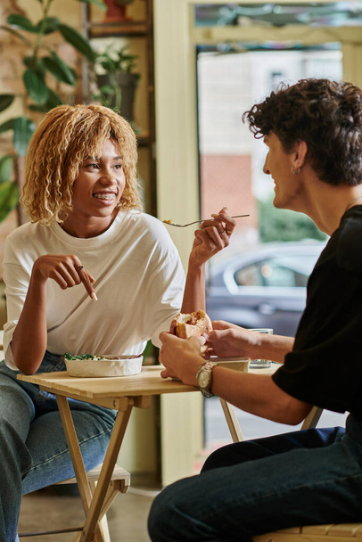 щаслива афроамериканка з брекетами, що їдять салат біля розмитого кучерявого хлопця у веганському кафе - Фото, зображення