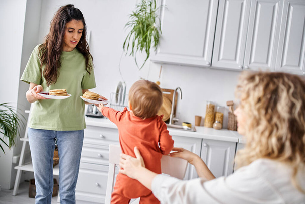joyful brunette woman sharing fresh pancakes with her blurred blonde partner and their baby girl - Photo, Image
