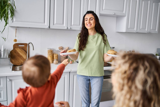 beautiful brunette woman sharing fresh pancakes with her blurred blonde partner and their baby girl - Photo, Image