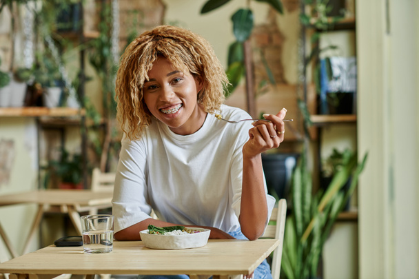 gelukkig Afrikaans amerikaans vrouw in beugel eten verse salade in kom in van plant gevulde veganistisch cafe - Foto, afbeelding