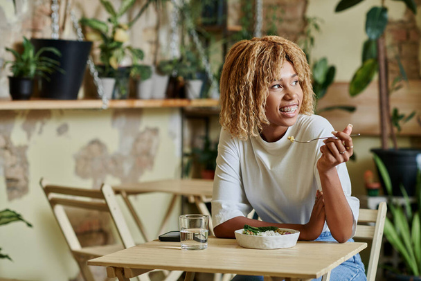 chica afroamericana feliz en frenillos comiendo ensalada fresca en un tazón dentro de la cafetería vegana llena de plantas - Foto, Imagen