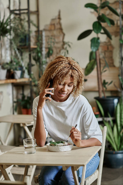 menina americana africana feliz em aparelho falando no smartphone e comer salada vegan fresco no café - Foto, Imagem