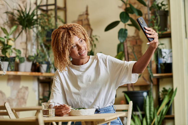 allegra ragazza afro-americana in apparecchio di prendere selfie mentre si mangia insalata vegana fresca nel caffè - Foto, immagini