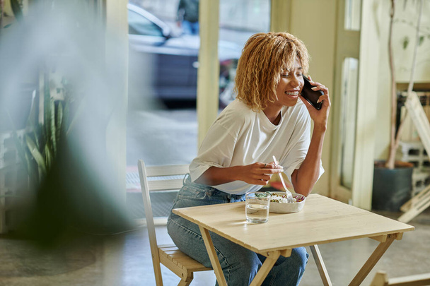 glückliche junge dunkelhäutige Frau in Hosenträgern, die mit dem Smartphone spricht und im Café frischen veganen Salat isst - Foto, Bild