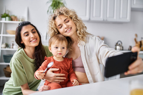 jolly beautiful lgbt couple taking selfie together with their daughter on kitchen, family concept - Photo, Image