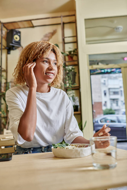 щаслива темношкіра молода жінка з брекетами, що тримає виделку біля свіжого веганського салату в сучасному кафе - Фото, зображення