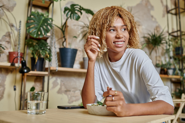 chica de piel oscura de ensueño con frenos tenedor cerca de ensaladera fresca en la cafetería vegana moderna - Foto, imagen