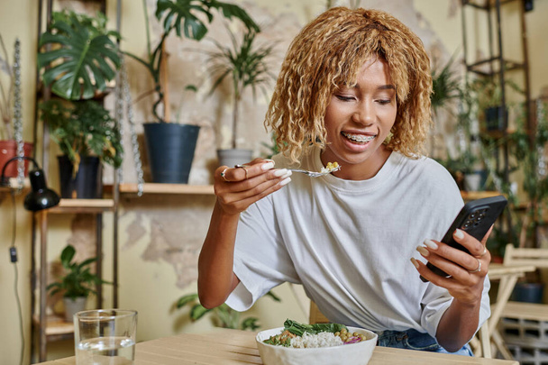 felice giovane donna dalla pelle scura con bretelle tenendo forchetta vicino insalatiera nel moderno caffè vegan - Foto, immagini