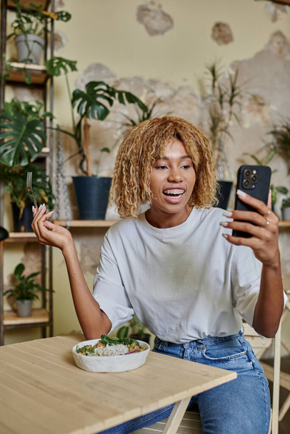 podekscytowany ciemnoskóry młoda kobieta z aparatem na zęby trzymając widelec w pobliżu sałatki wegańskiej i patrząc na smartfona - Zdjęcie, obraz