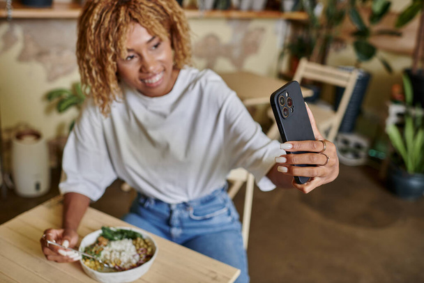 mujer afroamericana borrosa tomando selfie en el teléfono inteligente en la cafetería vegana llena de plantas, hora de la comida - Foto, imagen
