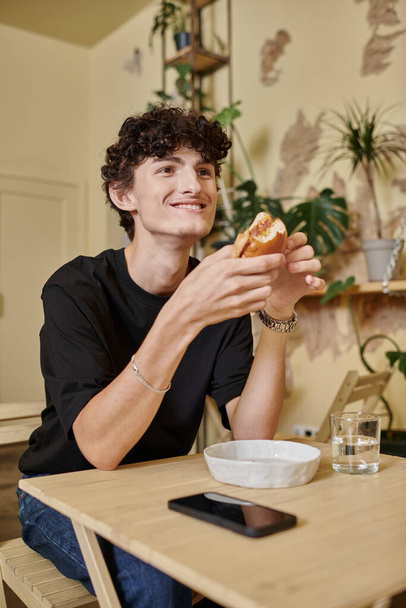 positive and curly young man holding organic tofu burger and smiling in plant filled vegan cafe - Fotoğraf, Görsel