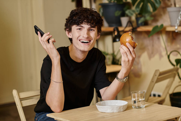 happy and curly young man holding plant-based tofu burger and smartphone in vegan cafe, city life - Photo, Image