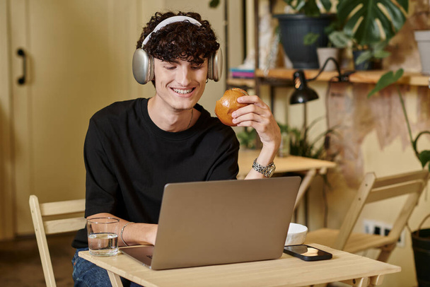веселый человек в беспроводных наушниках с помощью ноутбука и наслаждаясь тофу бургер в веганском кафе, завод на основе - Фото, изображение