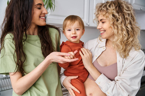 bonded cheerful lesbian couple in homewear posing happily with their baby girl, modern parenting - Photo, Image