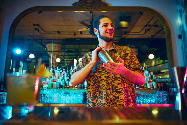 Vista dal basso rom bar bancone del giovane barista scuotendo attivamente cocktail shaker rendendo alcol dolce cocktail gustosi al bar con un design moderno. Concetto di tempo di festa, night club, stile di vita delle persone. - Foto, immagini