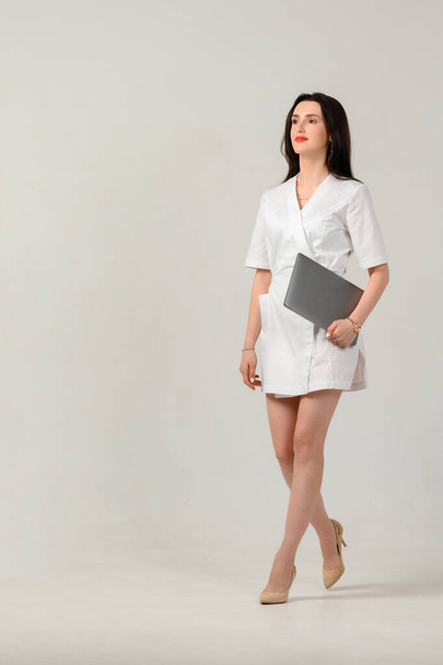 косметолог в белом халате держит в руках ноутбук в фотостудии на белом фоне, портрет девушки в белом халате. - Фото, изображение