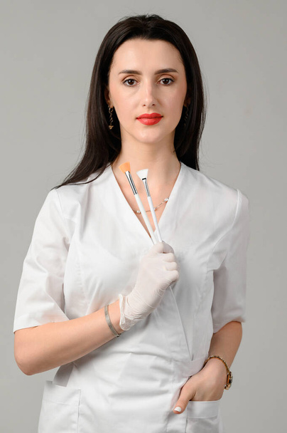 το κορίτσι cosmetologist κατέχει δύο βούρτσες στα χέρια της σε λευκό φόντο, το κορίτσι είναι ένας αισθητικός σε λευκά γάντια μιας χρήσης. - Φωτογραφία, εικόνα