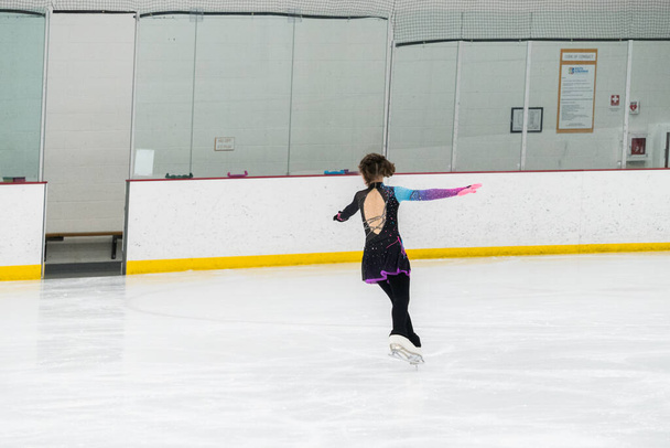 Jovencita perfeccionando su rutina de patinaje artístico mientras usa su vestido de competición en una pista de hielo interior. - Foto, Imagen