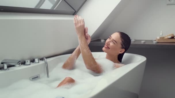 4K videobeelden, opgetogen vrouw ondergedompeld in schuim gevulde bad, zachtjes wassen van haar handen en speels bezig met bubbels. De scène vangt een mix van ontspanning en kinderlijke vreugde, slow motion - Video
