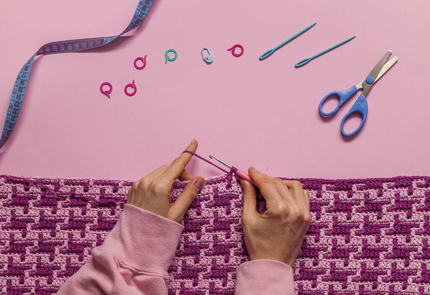Mãos de mulher crochê um cobertor de mosaico roxo. Tecido de crochê e ferramentas de crochê em um fundo rosa. Vista superior. Espaço de cópia. - Foto, Imagem