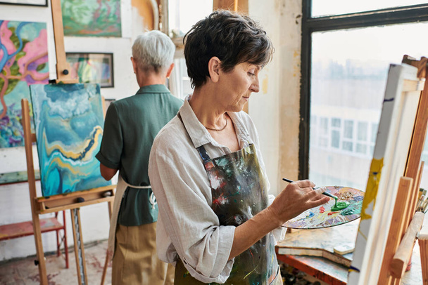 ταλαντούχα μελαχρινή ώριμη γυναίκα ζωγραφική στο καβαλέτο κοντά θηλυκό φίλο στο εργαστήριο τέχνης, δημιουργικό χόμπι - Φωτογραφία, εικόνα