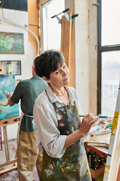 thoughtful brunette mature woman painting on easel near female friend in art workshop, creativity - Photo, Image