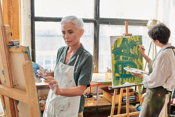 mujer madura de pelo gris en la pintura de delantal en caballete cerca de una amiga en el taller de arte, creatividad - Foto, imagen