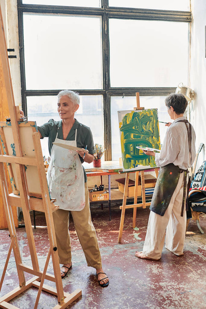 talentierte Frauen mittleren Alters in Schürzen malen auf Staffeleien in der Werkstatt für moderne Kunst, kreatives Hobby - Foto, Bild
