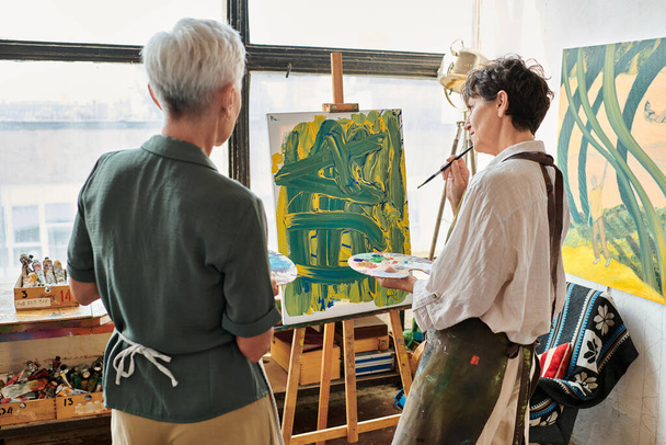 задумчивая зрелая женщина смотрит на свою картину возле подруги в мастерской ремесла, творческое хобби - Фото, изображение