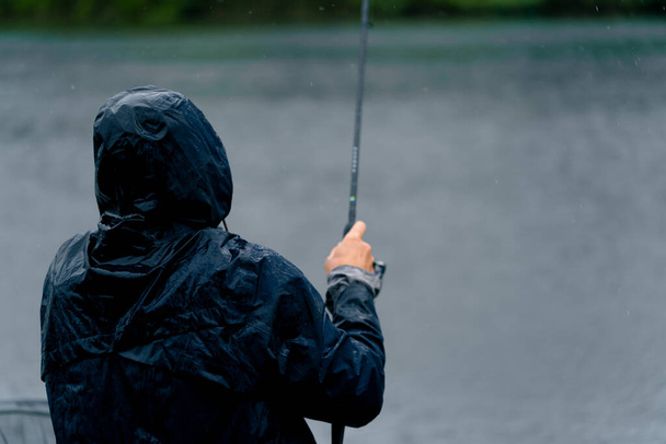 Pescador en impermeable con caña de pescar o herramientas profesionales de spinning sentado en la orilla del río vista trasera Tire de los peces de la pesca deportiva lago - Foto, Imagen