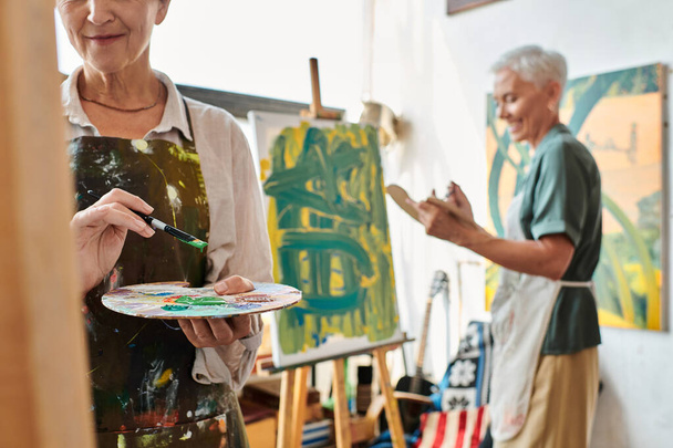 mujer madura con paleta cerca sonriente amiga pintura sobre caballete en taller de arte, creatividad - Foto, Imagen