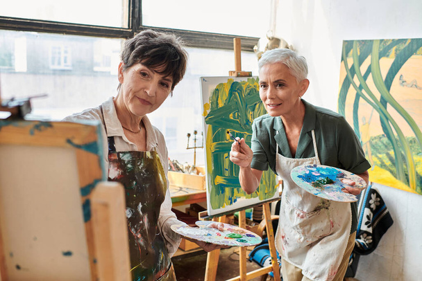 ώριμες γυναίκες καλλιτέχνες με παλέτες που κοιτάζουν το καβαλέτο στο εργαστήριο τέχνης, τη δημιουργικότητα και τη συνεργασία - Φωτογραφία, εικόνα