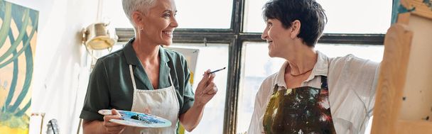 joyeuses artistes féminines matures dans des tabliers tenant des palettes et parlant dans un atelier d'art moderne, bannière - Photo, image