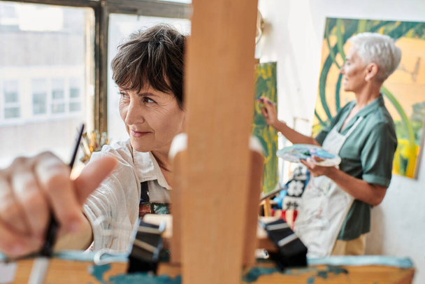kreativní zralá žena v blízkosti stojanu a přítelkyně malování na rozmazaném pozadí, umělecký koníček - Fotografie, Obrázek