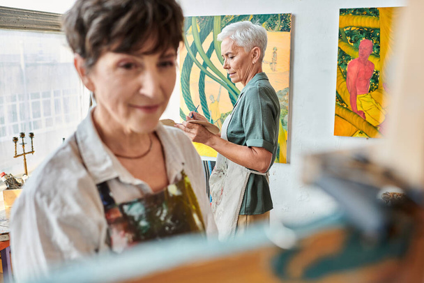 θολή γυναίκα καλλιτέχνης κοντά easel και ώριμη γυναίκα ζωγραφική στο εργαστήριο τέχνης, δημιουργικό χόμπι - Φωτογραφία, εικόνα