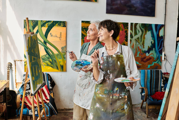 χαρούμενη κομψή ώριμες γυναίκες σε ποδιές κοιτάζοντας μακριά στο εργαστήριο σύγχρονης τέχνης, τη φιλία και τη δημιουργικότητα - Φωτογραφία, εικόνα