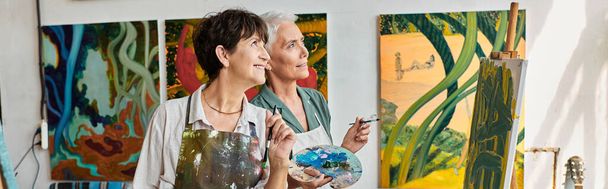 radosne kreatywne dojrzałe kobiety w fartuchach odwracając wzrok w pracowni sztuki nowoczesnej, baner poziomy - Zdjęcie, obraz