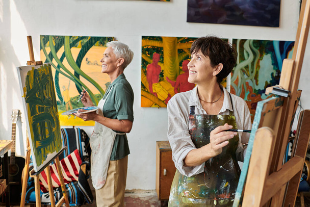 sonrientes mujeres maduras en delantales pintando en caballetes y mirando hacia otro lado en el taller de arte, hobby creativo - Foto, imagen
