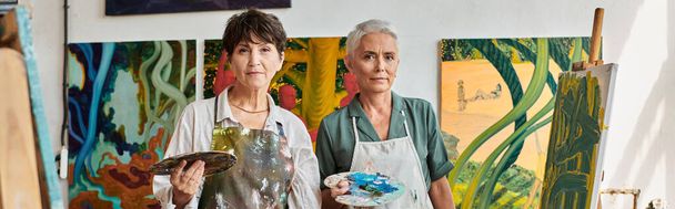 talentoso pintores do sexo feminino em aventais segurando paletas e olhando para a câmera no estúdio de arte, banner - Foto, Imagem