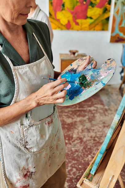 cropped view of mature female artist mixing on colors on palette with brush in art workshop - Φωτογραφία, εικόνα